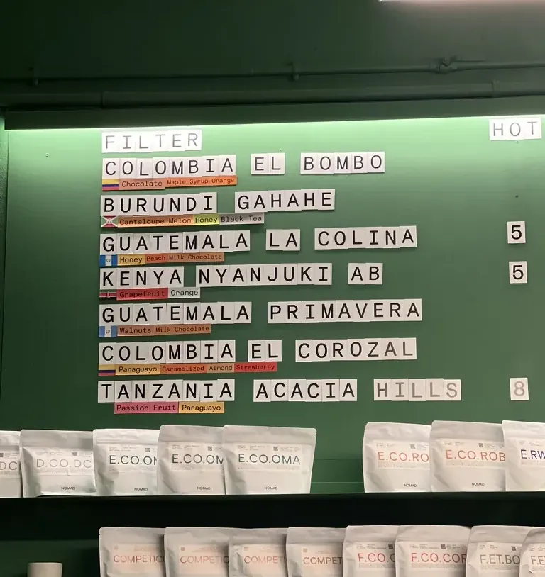 Quadro de cafés filtrados na parede verde da Nomad Coffee, com os pacotes embaixo