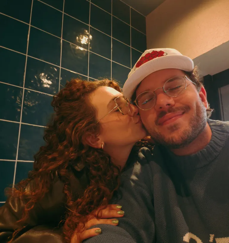 A namorada do Leo dando um beijo no rosto dele, numa parede de azulejos verdes
